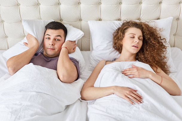 Stressful man suffers from insomnia, keeps pillow on head while hears wifes snoring during night, sleep together in one bed. Curly woman has problems with health, interrupts man to sleep well - Fotó, kép