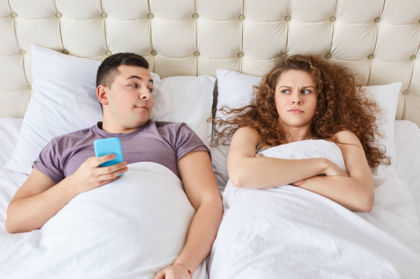 Addcited man holds modern cell phone, surfes in social networks, angry wife feels jealous, needs attention, stay together in bed, have problems with relationships. Bedtime and family concept - Photo, Image