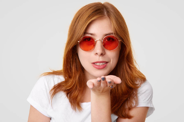 Headshot of coquette ginger female blows kiss at camera, expresses love to boyfriend, wears trendy round red sunglasses, dressed in casual t shirt, isolated over white background. Woman sends kiss - Fotoğraf, Görsel