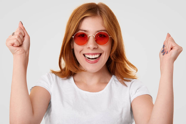 Overjoyed female student raises fists with triumph and happiness, rejoices successful passed exam, wears red sunglasses, poses against white background. People, positiveness, success concept - Photo, Image