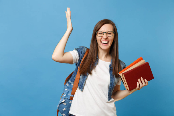 Портрет молодої смішної милої жінки студентки в окулярах з рюкзаком, що розтягує руку, тримає шкільні книги ізольовані на синьому фоні. Освіта в університетському коледжі концепція
 - Фото, зображення