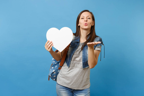 Portrait of young attractive cute woman student with backpack holding white heart with copy space sending air kisses isolated on blue background. Education in university. Copy space for advertisement - Photo, image