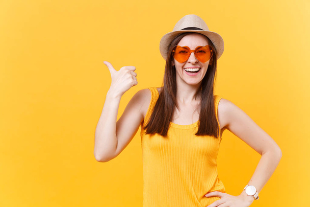 Πορτρέτο του χαμογελώντας νεαρή γυναίκα με Καπέλο αχύρου καλοκαίρι, πορτοκαλί γυαλιά δείχνει δεξιά στην άκρη αντίγραφο χώρου που απομονώνονται σε κίτρινο φόντο. Άνθρωποι ειλικρινή συναισθήματα, αντίληψη του τρόπου ζωής. Περιοχή διαφήμισης - Φωτογραφία, εικόνα