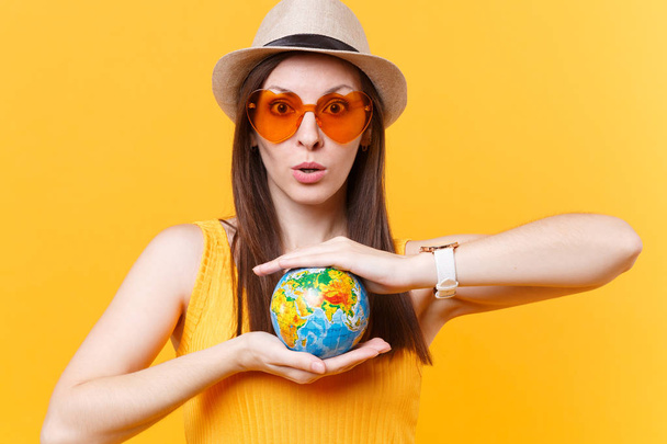 Turisztikai nyári kalap nő portréja, narancsszínű szemüveg tart kezében globe elszigetelt, a sárga háttér. Menekülés hétvégén utazni külföldre utazik. Légi járat út fogalma. Állítsa le a természet szemetet - Fotó, kép