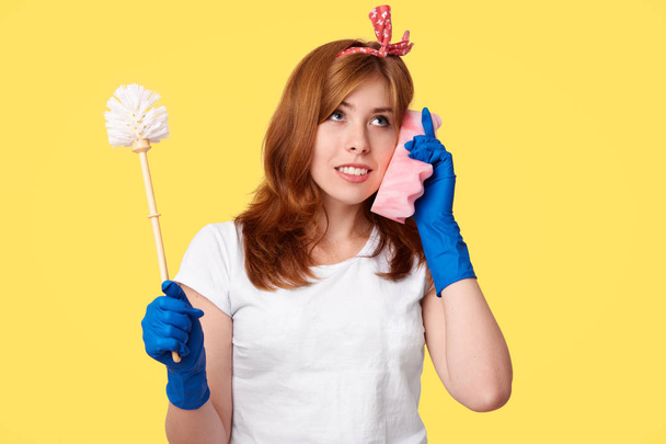 Studio shot of attentive housemaid holds brush, uses sponge as mobile phone, dicusses something with friend, dressed in casual clothes, poses against yellow background. Cleaning and hygiene concept - Photo, Image