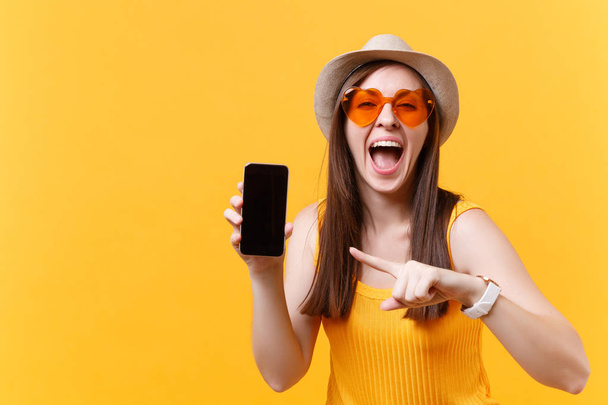 Portrait of excited amazed woman in summer hat, orange glasses keep mouth wide open, look surprised, hold mobile phone with blank empty screen isolated on yellow background. People emotions concept - Photo, Image