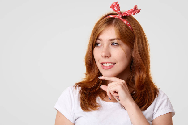 Happy Caucasian female with positive smile, holds chin and looks gladfully aside, wears stylish headband and white t shirt, poses against studio wall with copy space for your advertisement or text - Photo, Image