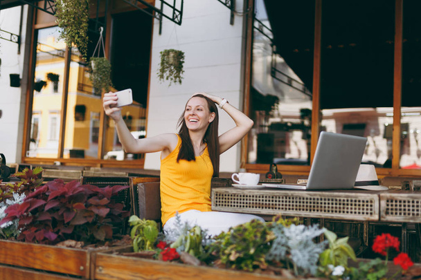 Ευτυχισμένος κορίτσι χαμογελαστό στην υπαίθρια καφέ καφέ μικροπωλητή κάθεται στο τραπέζι με φορητό υπολογιστή, κάνει λήψη selfie πυροβόλησε στο κινητό τηλέφωνο στο εστιατόριο κατά τη διάρκεια του ελεύθερου χρόνου. Κινητό γραφείο freelance έννοια - Φωτογραφία, εικόνα