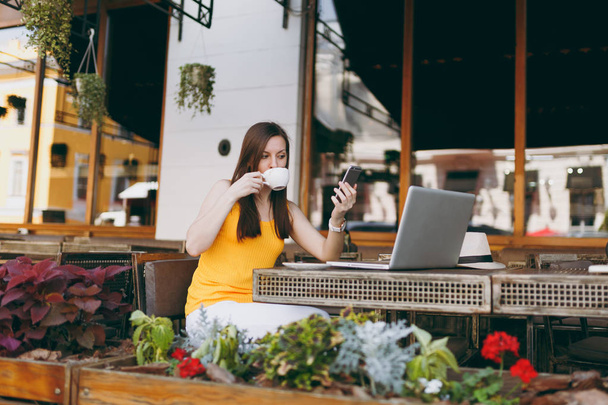 Szczęśliwa dziewczyna na zewnątrz ulicznych kawiarni Café siedzi w tabeli z komputerem pc laptop, wiadomość SMS na telefon komórkowy, pić herbatę cup w restauracji w wolnym czasie. Mobilne biuro tłumaczy koncepcję - Zdjęcie, obraz
