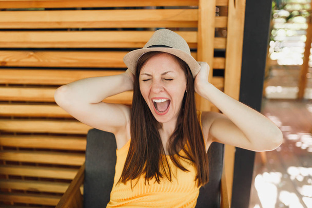 Porträt einer aufgeregt lächelnden jungen Frau mit Strohhut und gelbem Hemd, die die Hände auf den Kopf gelegt hat, auf hölzernem Hintergrund im Sommer-Café auf der Straße. Menschen aufrichtige Emotionen, Lebensstil-Konzept - Foto, Bild