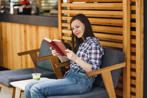 Piękna kobieta w na zewnątrz ulicy lato kawiarnia café drewniane siedzi w ubranie, czytając książkę z filiżanka koktajl, relaks restauracja w wolnym czasie. Styl życia odpoczynek wakacje koncepcja - Zdjęcie, obraz