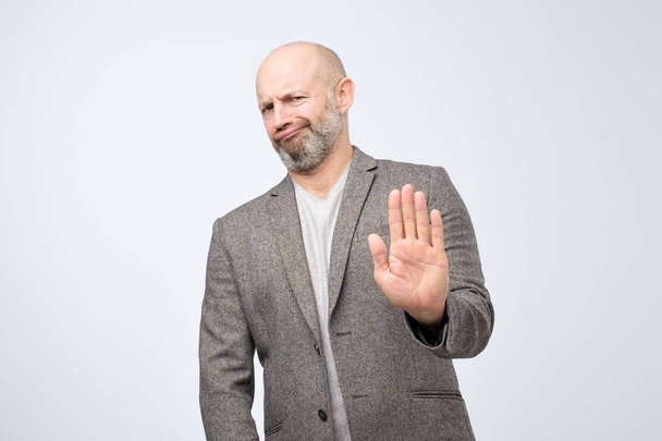 Attractive mature man showing refusal gesture. It is not for me, leave me in piece, has angry expression, poses against white concrete studio background - Photo, Image