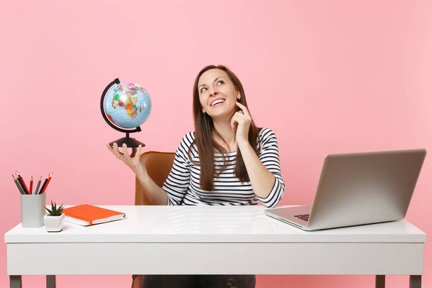 Young pensive woman holding globe, dreaming planning vacation while sit, work at office with contemporary pc laptop isolated on pastel pink background. Achievement business career concept. Copy space - Foto, immagini
