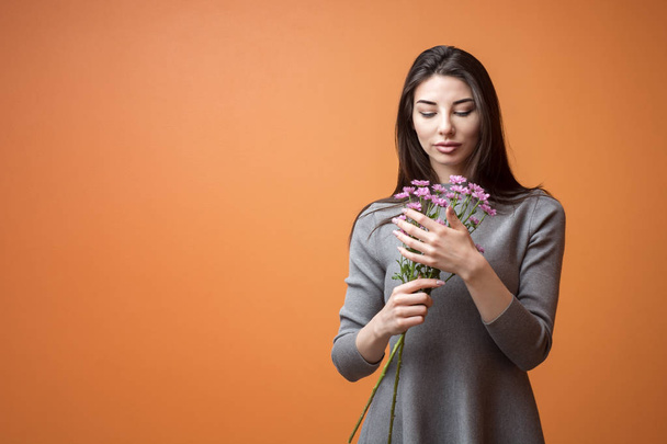 Portrait of a young beautiful brunette woman in grey dress holding violet flowers in her hands and looking at it against orange background - Photo, Image