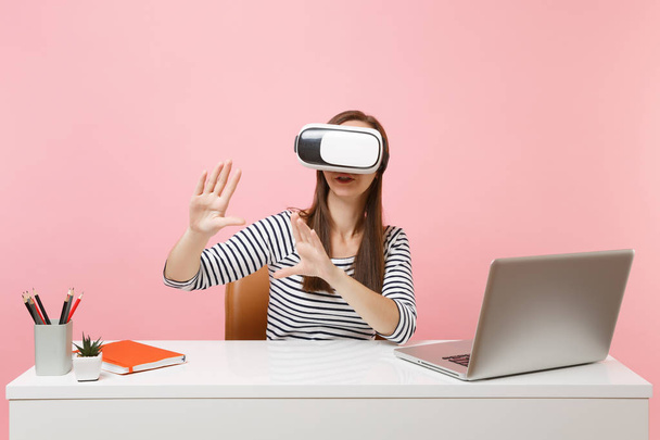 Girl in headset of virtual reality on head touch something like push on button or pointing at floating virtual screen work at desk with laptop isolated on pink background. Achievement business career - Foto, afbeelding