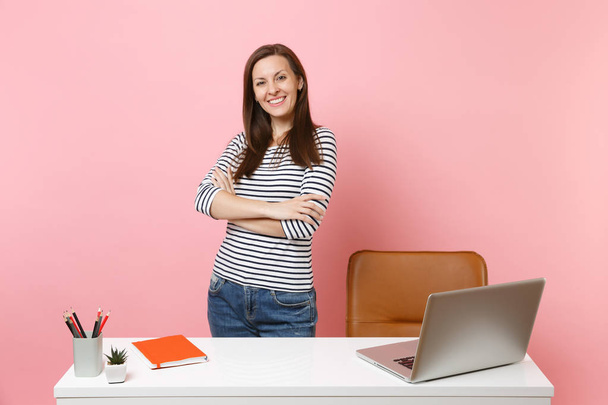 Mladá úspěšná dívka v neformálním oblečení práce stojící poblíž bílý stůl s moderním pc laptop izolované na pastelově růžové pozadí. Úspěch obchodní kariéru koncept. Kopírovat prostor pro reklamu - Fotografie, Obrázek
