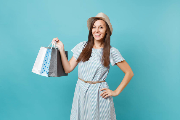 Portret van modieuze lachende Kaukasische vrouw in zomer jurk, stro hoed bedrijf pakketten zakken met aankopen na winkelen geïsoleerd op blauwe pastel achtergrond. Ruimte voor reclame kopiëren. - Foto, afbeelding