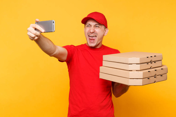 Consegna uomo in rosso cap t-shirt dando cibo ordinare pizza italiana in scatole flatbox vuote su sfondo giallo. Uomo dipendente pizzaman corriere facendo scattare selfie sparato sul cellulare. Concetto di servizio
 - Foto, immagini