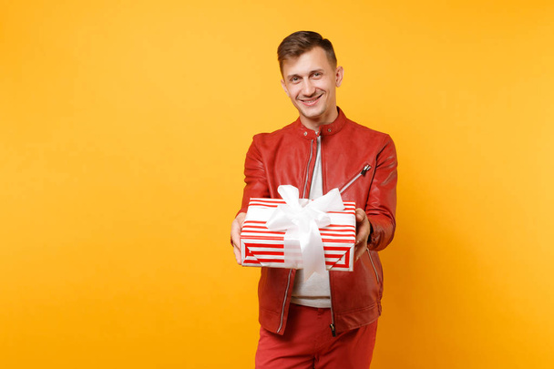 Ritratto voga sorridente bel giovane uomo in giacca rossa, t-shirt tenere presente scatola con regalo isolato su sfondo giallo trend luminoso. Persone emozioni sincere concetto di stile di vita. Area pubblicitaria
 - Foto, immagini