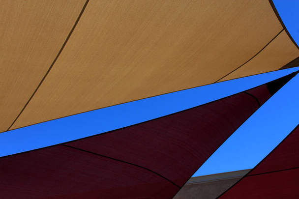 farbenfrohe abstrakte geometrische Designs, die durch Planen geschaffen wurden, die für den Schutz vor der Sonne im Freien verwendet werden und verschiedene Arten von Winkeln zeigen, zusammen mit einem sattblauen Hintergrund des klaren Himmels. - Foto, Bild