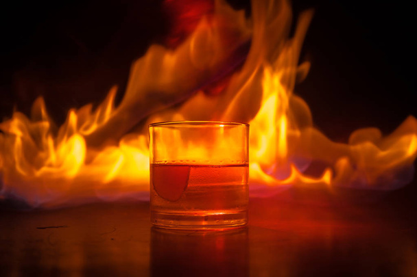 Whiskey in fire concept. Glass of whiskey and ice on wooden surface with color light and fog on background. Close up. Selective focus - Fotoğraf, Görsel