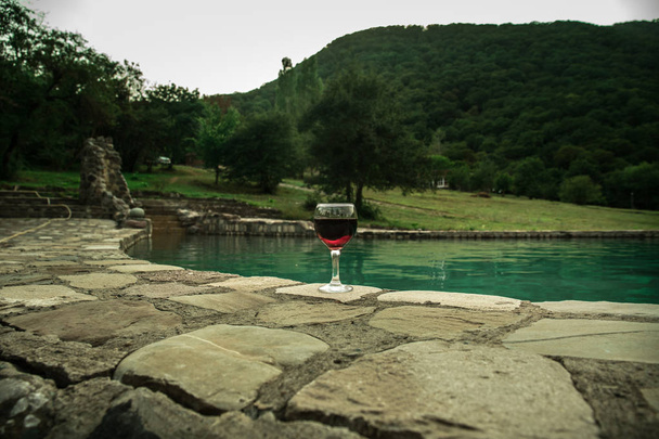 Drink in hoog glas in zwembad. Verfrissing op zomerdag. Paarse juice cocktail of vine. Berg bos achtergrond. Selectieve aandacht - Foto, afbeelding