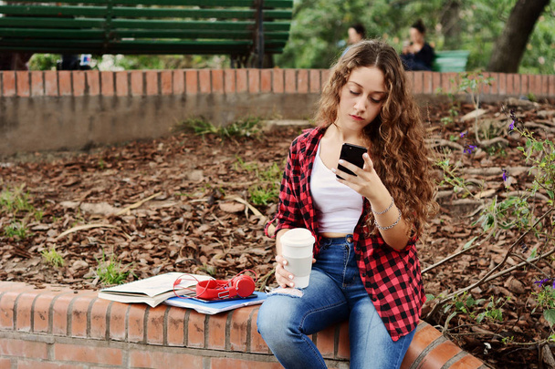 Chica estudiante joven con una tableta y una taza de café desechable sentado en el parque en otoño
. - Foto, Imagen