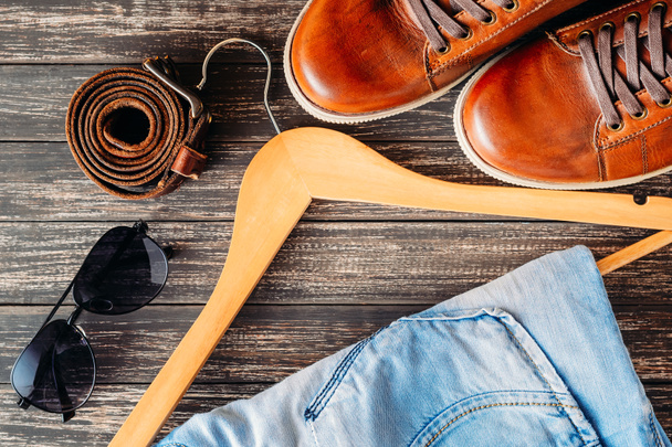 braune lederne Freizeitschuhe Jeans auf Kleiderbügel, Gürtel und Sonnenbrille auf Holzhintergrund Draufsicht - Foto, Bild