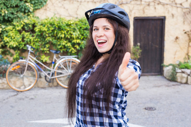 Молодая улыбающаяся женщина в шлеме и солнцезащитных очках показывает большие пальцы на заднем плане аллеи и велосипеде
 - Фото, изображение