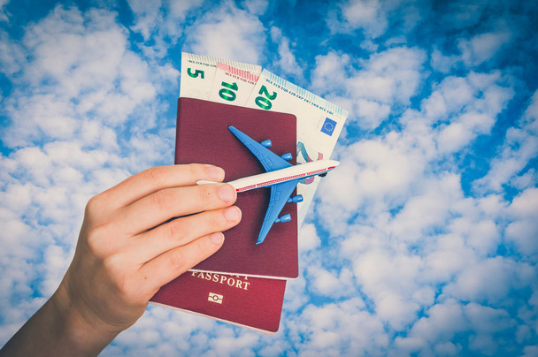 Avião, passaporte e dinheiro na mão feminina sobre fundo céu azul - conceito de viagem - estilo retro
 - Foto, Imagem