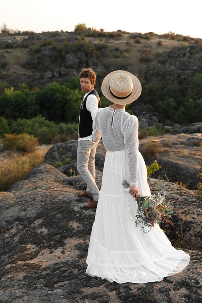 Happy newlyweds with beautiful field bouquet standing on rock outdoors - Zdjęcie, obraz