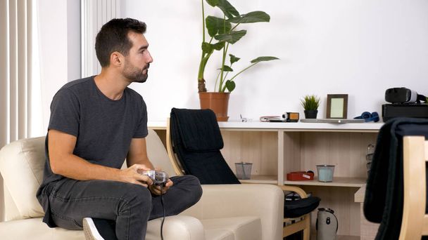 Привлекательный молодой человек, сидящий на диване и играющий в видеоигры
 - Фото, изображение