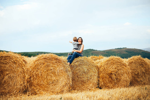 Мама и сын на закате сидят в поле на стоге сена и обнимаются под заходящим солнцем. Сельская жизнь, прогулка на природе, отдых в сельской местности
 - Фото, изображение