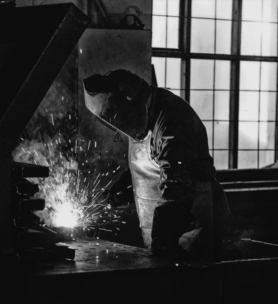 Welder at the factory in a welding mask welds metal parts, welding and sparks, black and white, welding holder - Foto, immagini