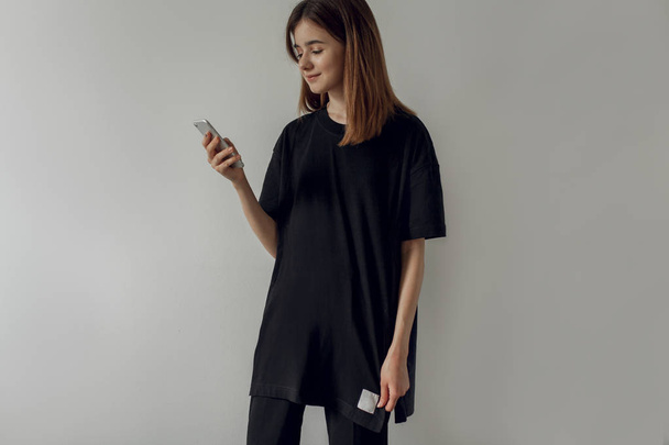 Photo of a smiling happy hipster girl wearing a casual black t-shirt using a mobile smartphone while standing on a white wall background at home. Young woman typing via cellphone - Photo, image