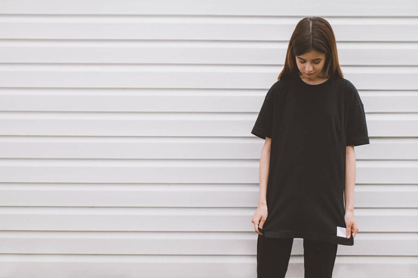 Photo of a girl with brown hair wearing a blank black t-shirt standing on a street wall background. Empty space for text or design. - Foto, Bild