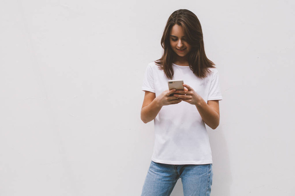 Smiling happy hipster girl wearing a white t-shirt using a mobile smartphone while standing on a white wall background. Young woman writing emails via cellphone - Foto, Bild