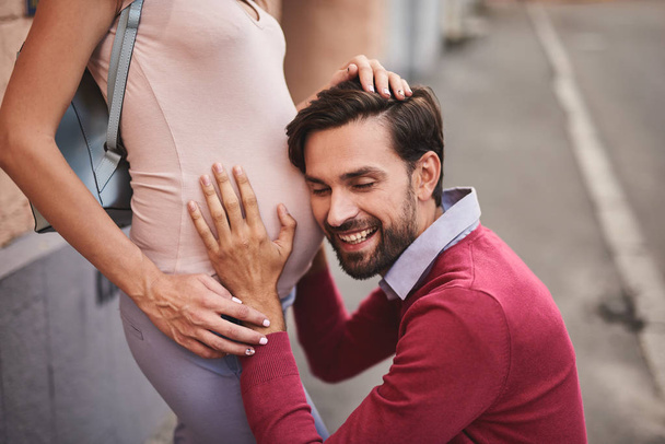 Smiling bearded man listening to tummy of his beautiful pregnant wife - Фото, изображение