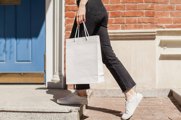 Young hipster smiling girl with white blank paper bag wearing white t-shirt and black jeans, mock-up of blank bag, brick wall and blue door on the background, concept of shopping - Fotoğraf, Görsel