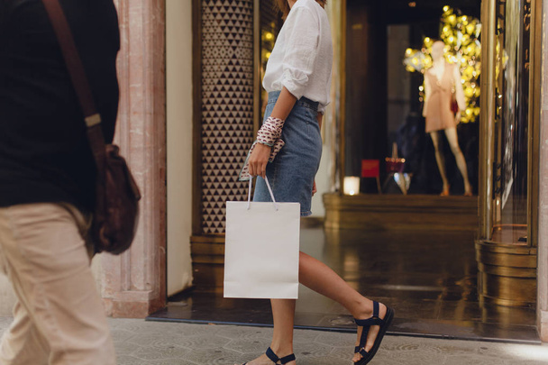 Cropped picture of young girl walking with white paper shopping bag, and doing shopping walking along the city streets. Concept of consumerism and shopping lifestyle - Фото, зображення