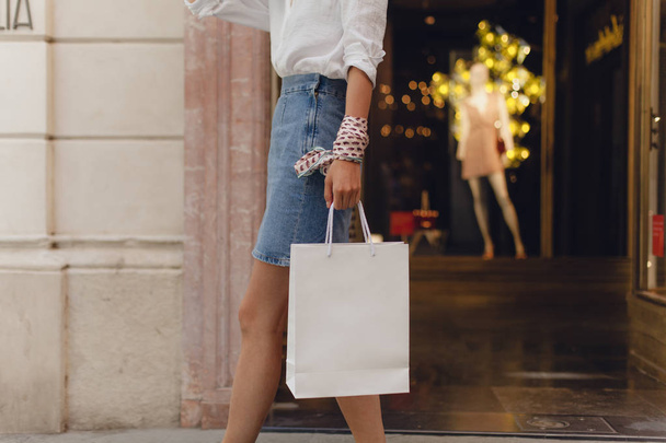 Cropped picture of young girl walking with white paper shopping bag and doing shopping walking along the city streets. Mockup of white blank shopping bag. Warm colors - Fotó, kép