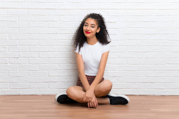 chica joven con el pelo rizado sentado en el suelo en una pared de ladrillo blanco
 - Foto, imagen