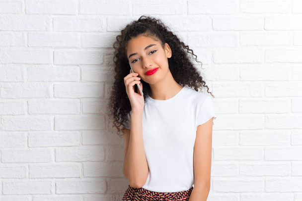young girl with curly hair using mobile phone on a white brick wall - Photo, Image