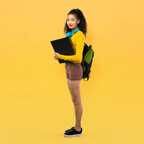 Corpo inteiro de menina estudante adolescente com cabelos encaracolados mantendo os braços cruzados em posição lateral enquanto sorri. Expressão confiante no fundo amarelo
 - Foto, Imagem