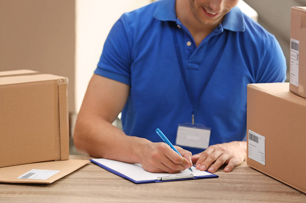 Young courier working with papers among parcels at table in delivery department - Foto, imagen