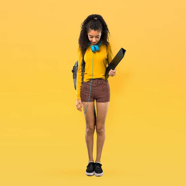 Corpo inteiro de adolescente estudante menina com cabelos cacheados de pé e olhando para baixo no fundo amarelo
 - Foto, Imagem