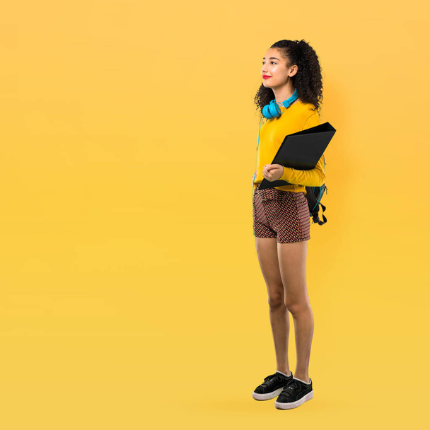 Πλήρες σώμα της έφηβος κορίτσι φοιτητής με σγουρά μαλλιά στέκεται και κοιτάζει προς την πλευρά σε κίτρινο φόντο - Φωτογραφία, εικόνα