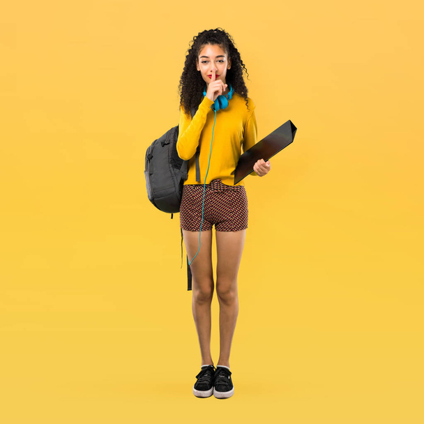 Cuerpo completo de adolescente estudiante con el pelo rizado que muestra un signo de cierre de la boca y el gesto de silencio poniendo el dedo en la boca sobre el fondo amarillo
 - Foto, imagen