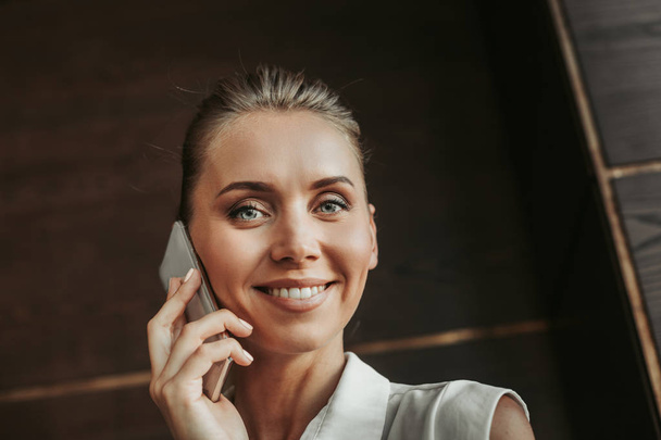Portrait of cheerful pretty female employer with attractive smile speaking on mobile while looking at camera inside. Pleased girl telling on gadget concept - Фото, изображение