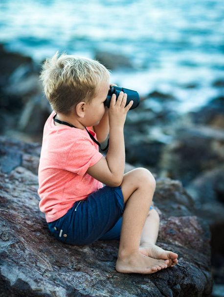 Little boy looking far away with binoculars, sitting on a rock near a sea - Фото, изображение